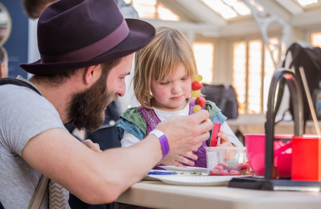 man in hat with small child holding fruit skewer